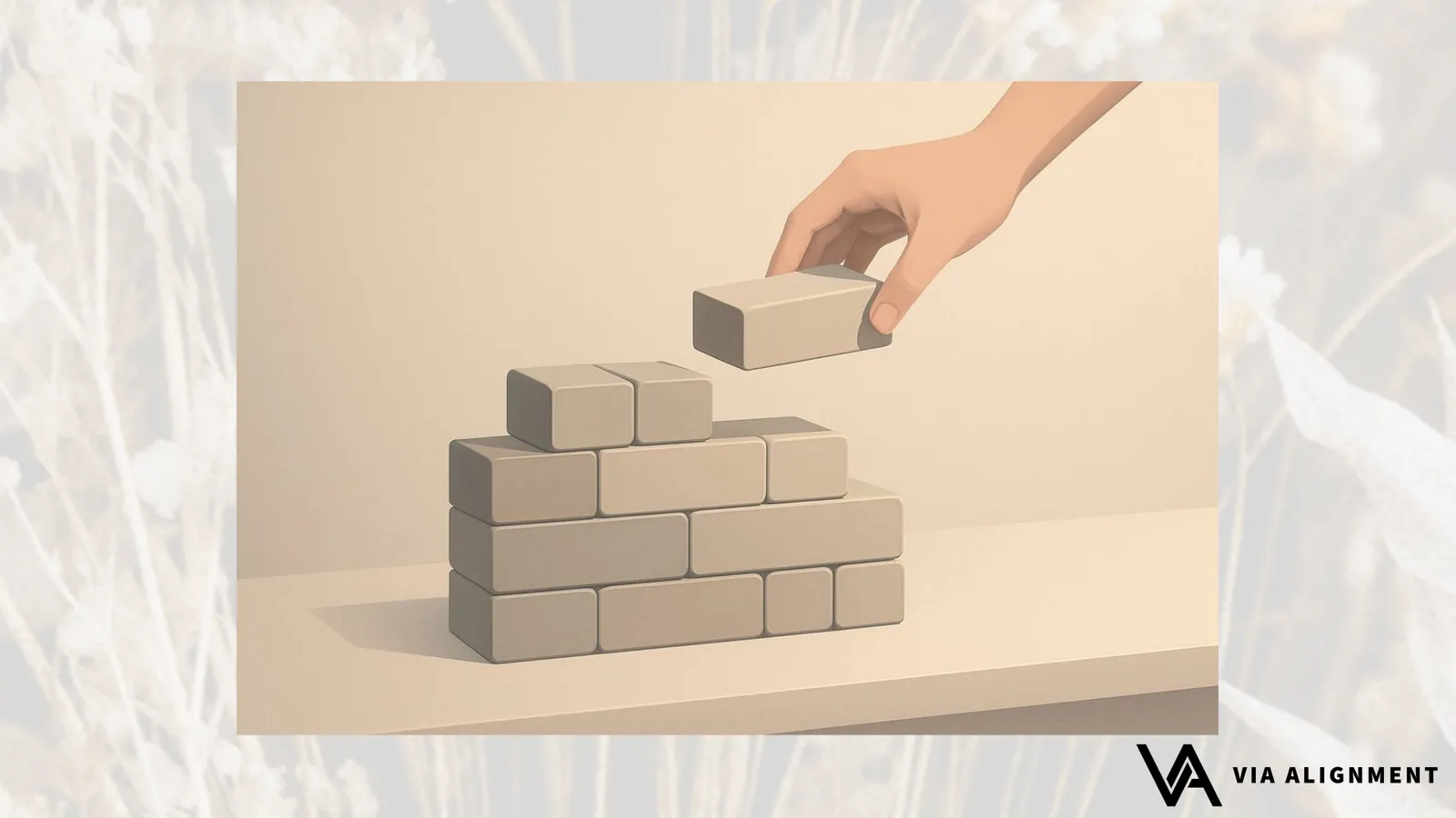 A hand places a small brick block onto the top of a stepped pyramid built from identical bricks on a tabletop.