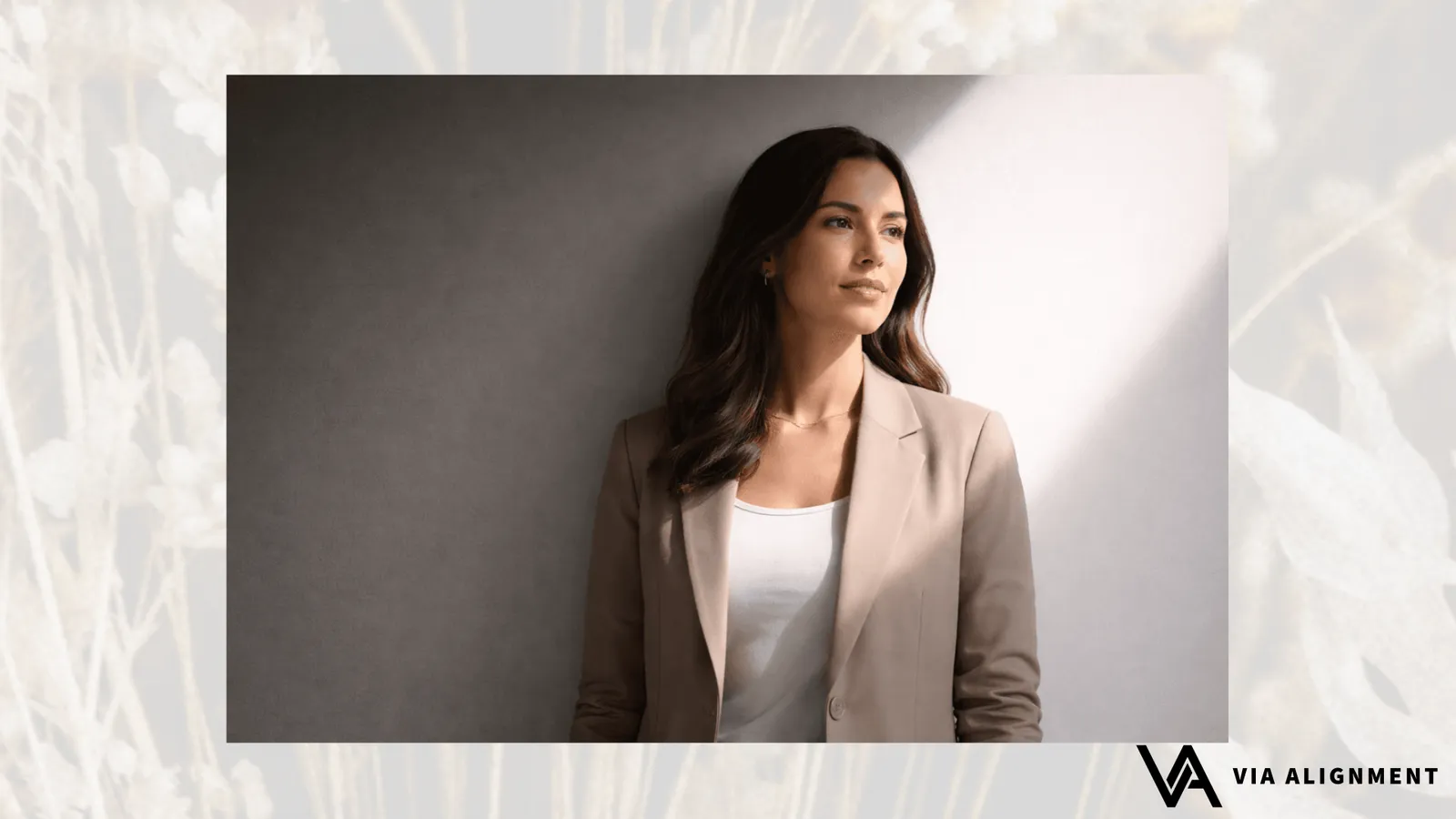 A woman in a tan blazer leans against a wall half in shadow and half in warm light, gazing calmly off to the side.